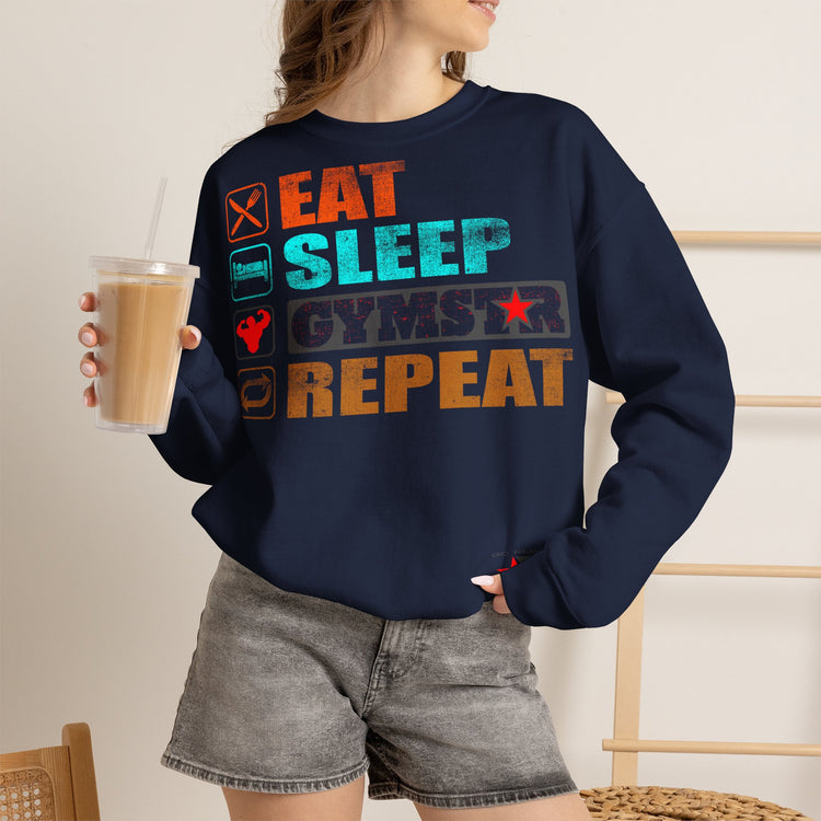 A woman wearing a black crewneck sweatshirt with a colorful graphic that reads 'EAT SLEEP GYMSTAR REPEAT' with fitness-themed icons, holding a iced coffee in a cafe setting.