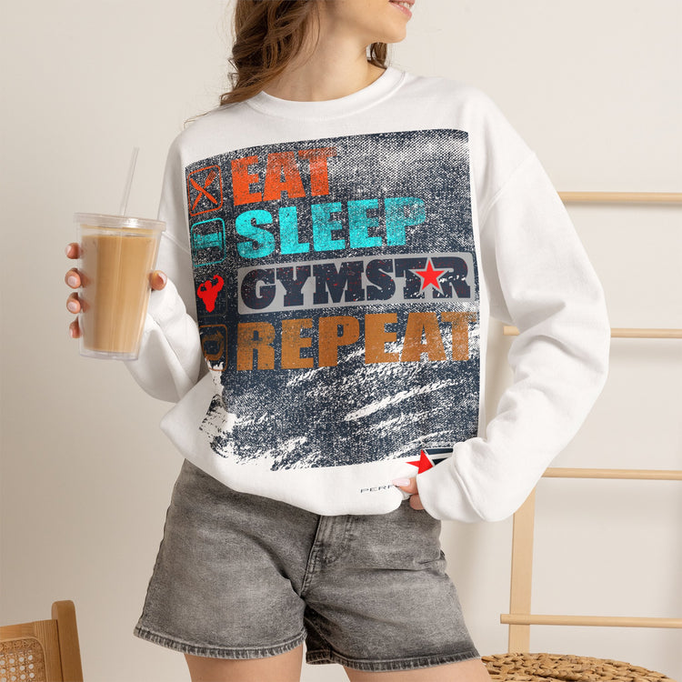 A woman wearing a white crewneck sweatshirt with a colorful distressed graphic that reads 'EAT SLEEP GYMSTAR REPEAT' in orange, teal, and dark blue. She is holding a cup of iced coffee and wearing gray denim shorts.