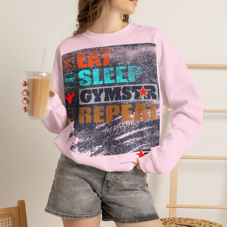 A woman wearing a light pink crewneck sweatshirt with a distressed graphic that reads 'EAT SLEEP GYMSTAR REPEAT' in orange, teal, and dark blue. She is holding a cup of iced coffee and wearing gray denim shorts.