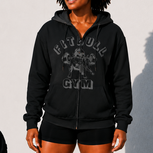 Person wearing a black 'Fitbull Gym' hoodie and shorts, standing next to gym equipment on a light background