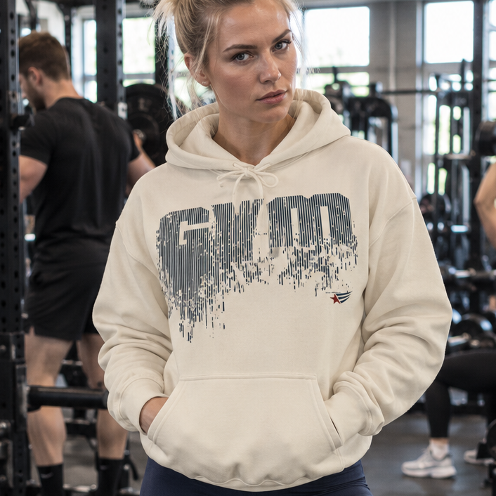 Emelie Johansson wearing a sand-colored GYMSTAR hoodie with a distressed graphic logo and dark navy leggings, posing in a modern fitness gym with athletes training in the background.