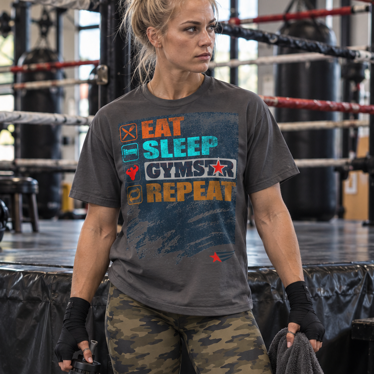 GYMSTAR model Emelie Johansson wearing a charcoal Eat Sleep GYMSTAR Repeat T-shirt and camouflage leggings, holding a water bottle and towel beside a boxing ring.