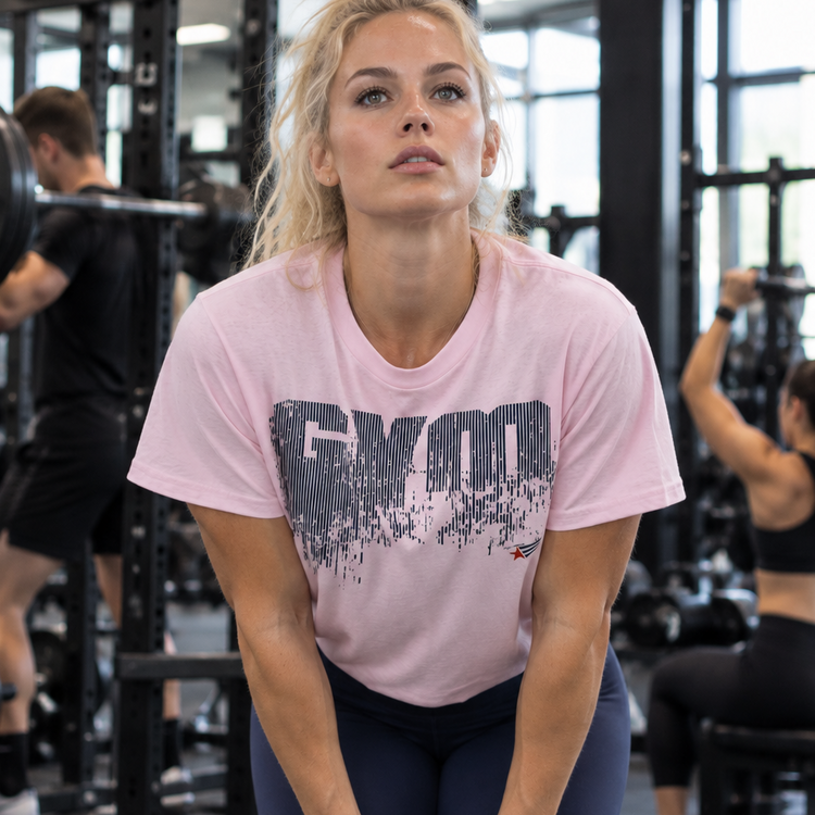 Emelie Johansson wearing a light pink GYMSTAR distressed logo T-shirt and navy blue leggings, leaning forward with hands on knees in a gym.