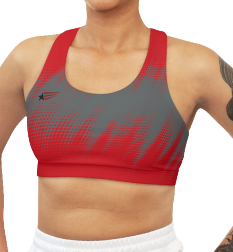 A woman wearing athletic sports bra, shown from the front. The main body is gray with a red, dotted, gradient pattern that radiates from the sides and bottom. The straps are solid red and form a racerback style at the back. A small logo featuring a star and three red and blue stripes is on the left side of the chest.