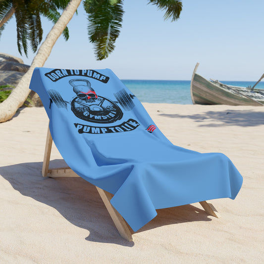 Light blue beach towel draped over a wooden lounge chair on sandy beach, featuring a black kettlebell and barbell graphic with a red skull, and the motivational text “BORN TO PUMP, PUMP TO LIVE,” by GYMSTAR.