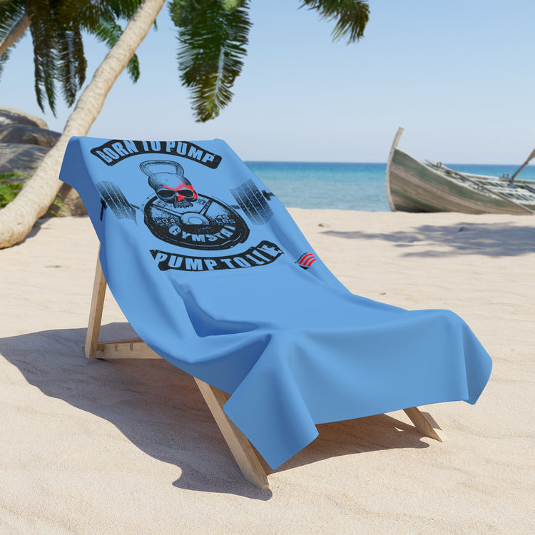 Light blue beach towel draped over a wooden lounge chair on sandy beach, featuring a black kettlebell and barbell graphic with a red skull, and the motivational text “BORN TO PUMP, PUMP TO LIVE,” by GYMSTAR.