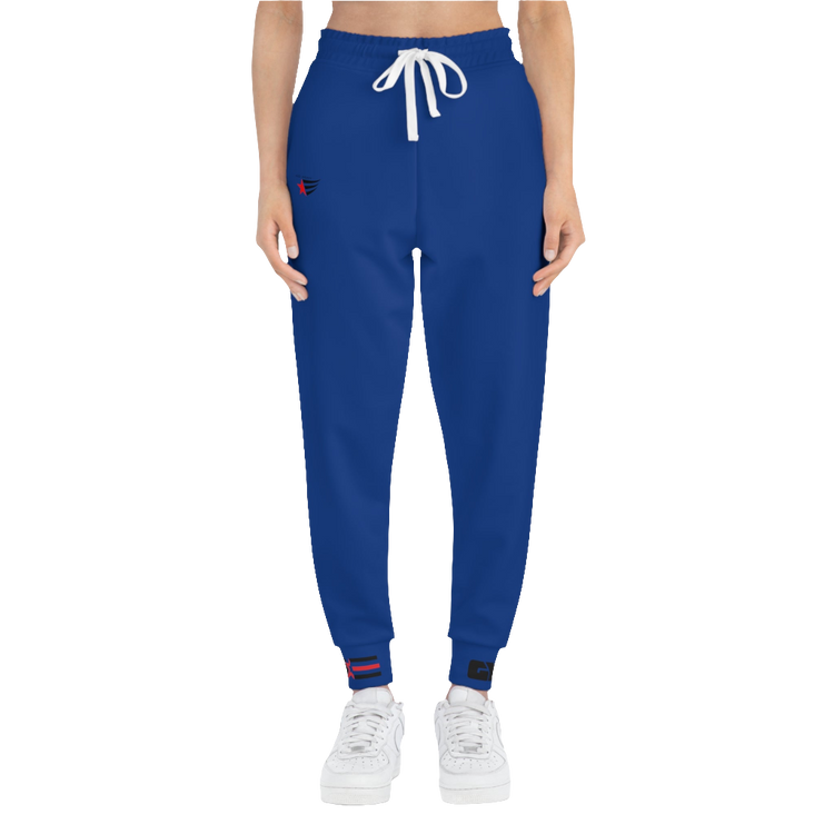 A woman wearing blue sports joggers with an elastic waistband and drawstrings, featuring a logo on the left leg and a white sneaker front side view.