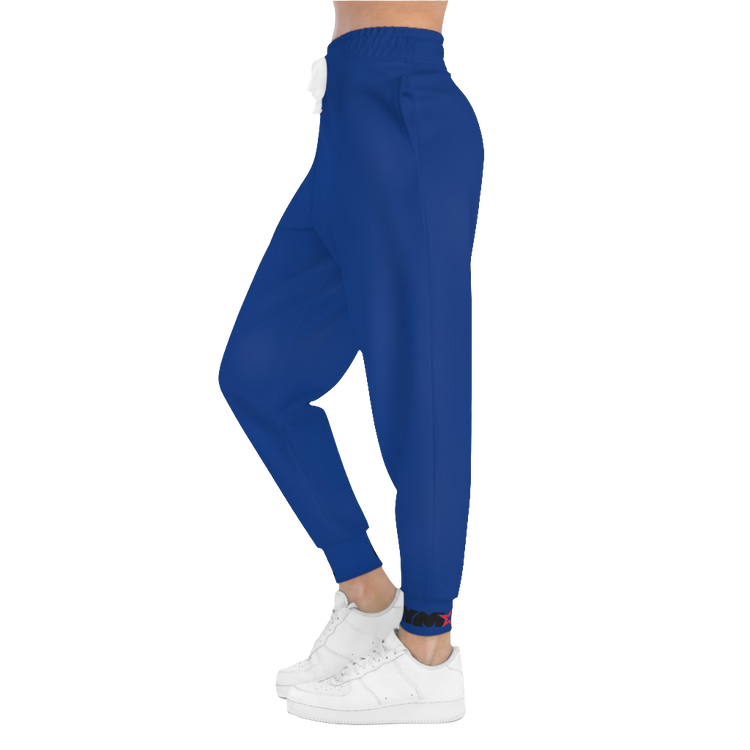 A woman wearing blue sports joggers with an elastic waistband and drawstrings, featuring a logo on the left leg and a white sneaker left side view.