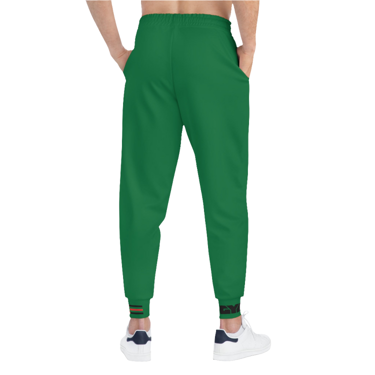 A person wearing green sports joggers with an elastic waistband and drawstrings, featuring a logo on the left leg and a white sneaker back side vew.