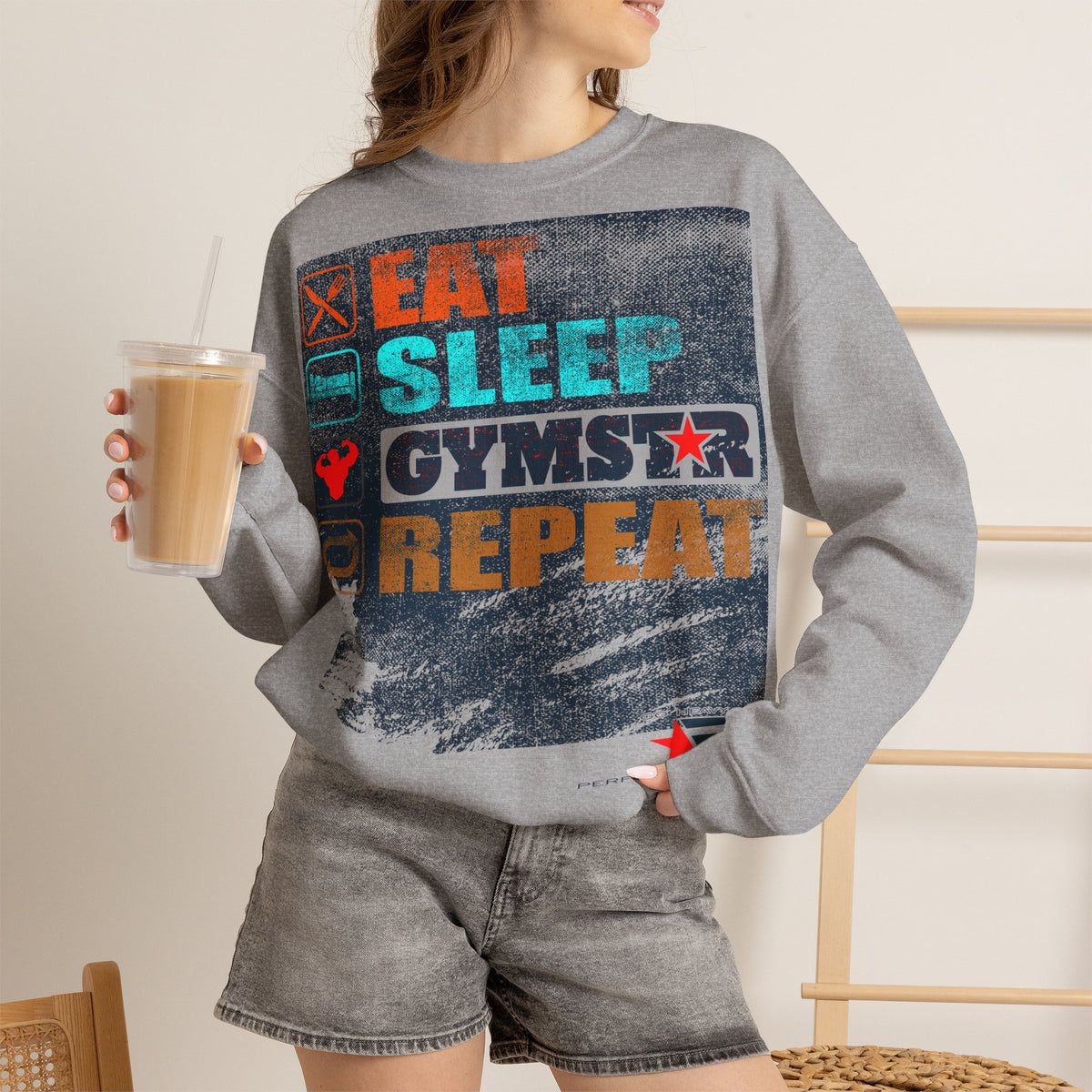 A woman wearing a heather gray crewneck sweatshirt with a distressed graphic that reads 'EAT SLEEP GYMS REPEAT' in orange, teal, and dark blue, paired with matching gray denim shorts. She is holding a cup of iced coffee in a brightly lit indoor setting.