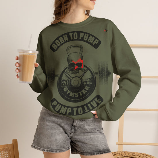A woman wearing an olive green crewneck sweatshirt with a black and red gym-themed graphic that reads 'BORN TO PUMP' and 'PUMP TO LIVE'. She is holding a cup of iced coffee in a bright, modern room.
