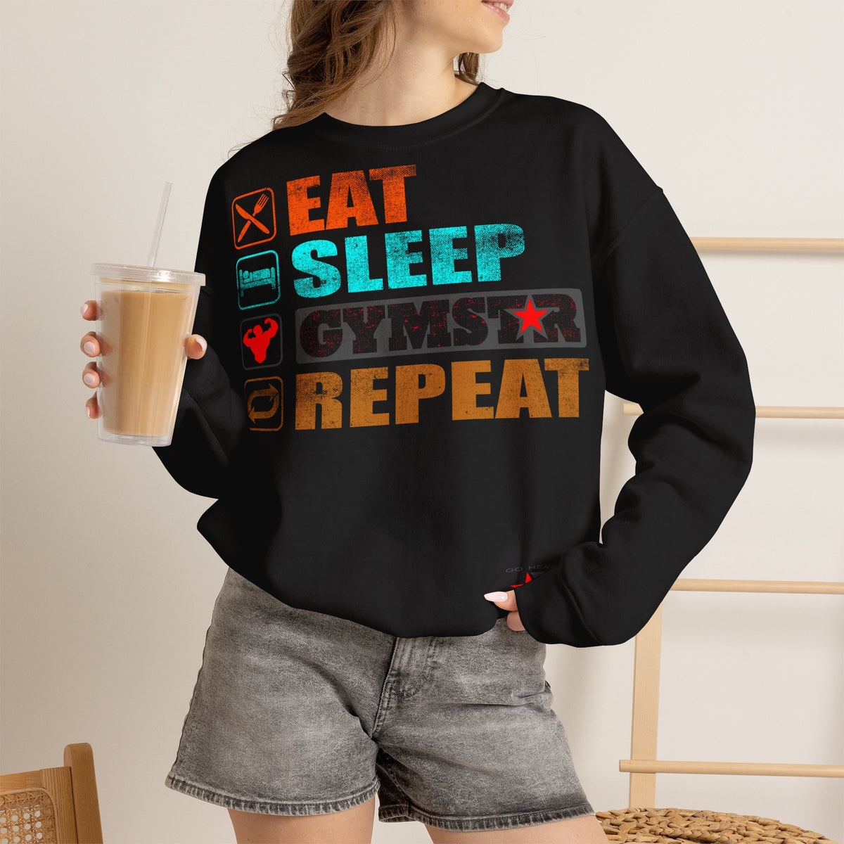A woman wearing a black crewneck sweatshirt with a colorful graphic that reads 'EAT SLEEP GYMSTAR REPEAT' with fitness-themed icons, holding a iced coffee in a cafe setting.