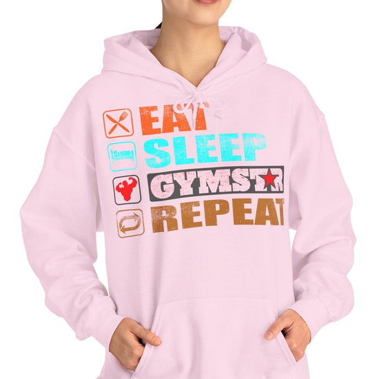 A woman modeling a light pink hoodie with a colorful graphic text design that reads 'EAT SLEEP GYMSTAR REPEAT' on a plain white background.