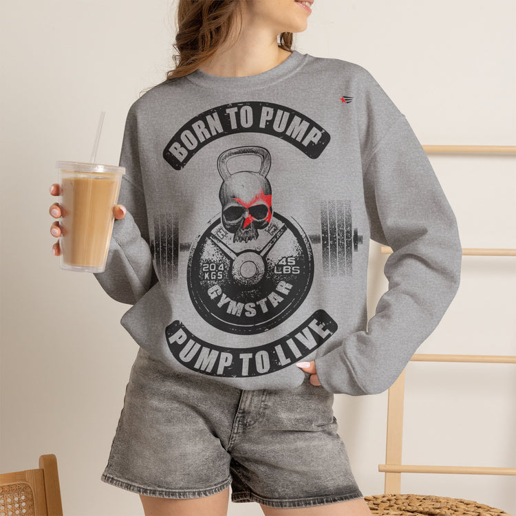 A woman wearing a heather gray crewneck sweatshirt with a large black and red graphic on the front that reads 'BORN TO PUMP' and 'PUMP TO LIVE' around a kettlebell and barbell design. She is holding an iced coffee and wearing gray denim shorts in a brightly lit indoor setting.