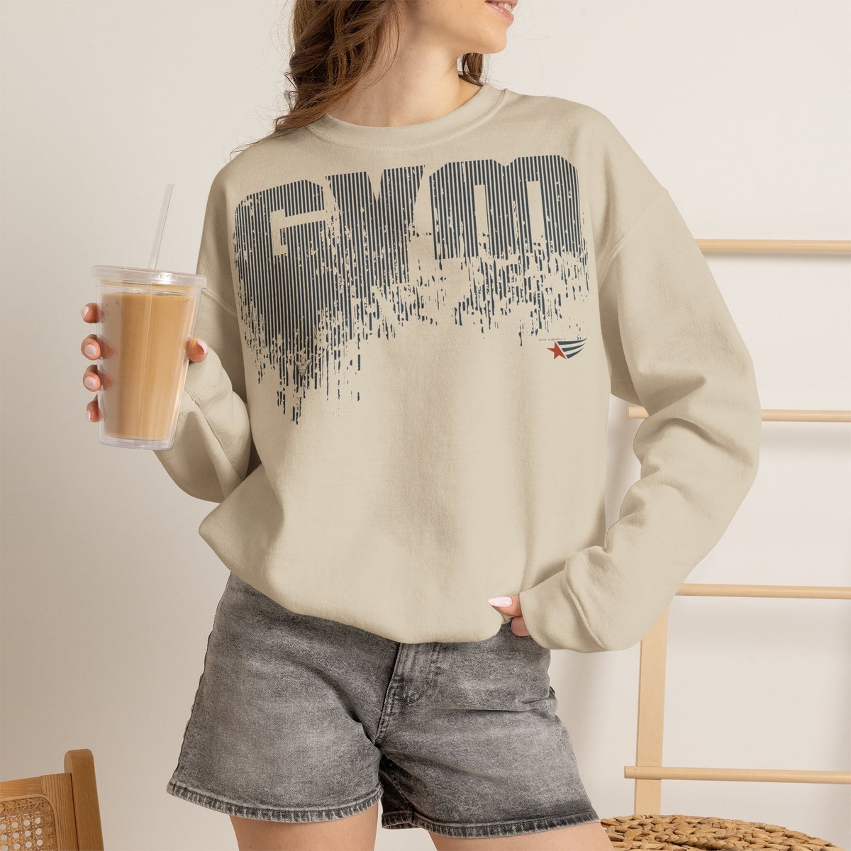 A woman wearing a beige crewneck sweatshirt with a dark gray distressed 'GYM' graphic on the front, paired with gray denim shorts and holding an iced coffee.