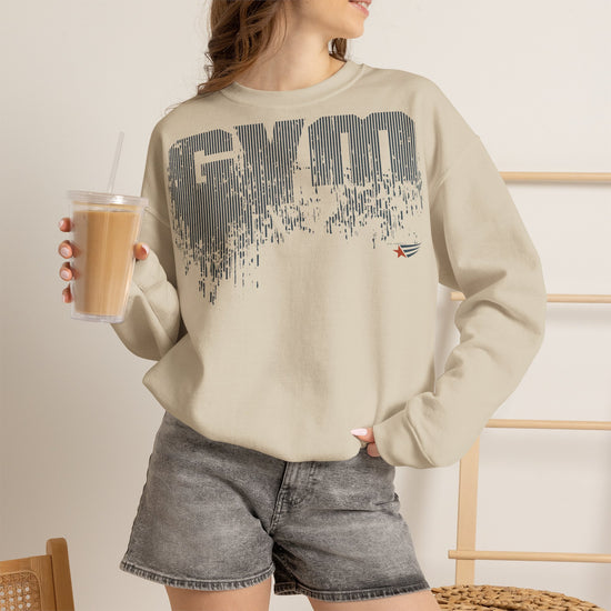 A woman wearing a beige crewneck sweatshirt with a dark gray distressed 'GYM' graphic on the front, paired with gray denim shorts and holding an iced coffee.