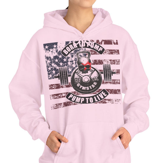 A woman modeling a light pink pullover hoodie with a large front graphic featuring a skull, barbell, American flag elements, and the words 'Rock the Pump Pump to Live' on a plain white background.