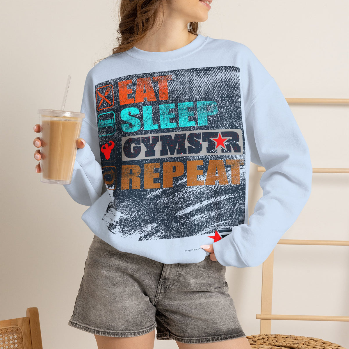 A woman wearing a light blue crewneck sweatshirt with a distressed graphic that reads 'EAT SLEEP GYMST REPEAT' in orange, teal, and black, paired with gray denim shorts and holding an iced coffee.
