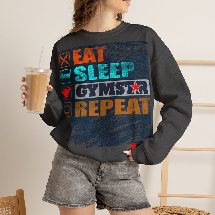 A woman wearing a dark charcoal gray sweatshirt with a colorful graphic that reads 'EAT SLEEP GYMSTER REPEAT' in orange, teal, and white, paired with gray denim shorts and holding an iced coffee.