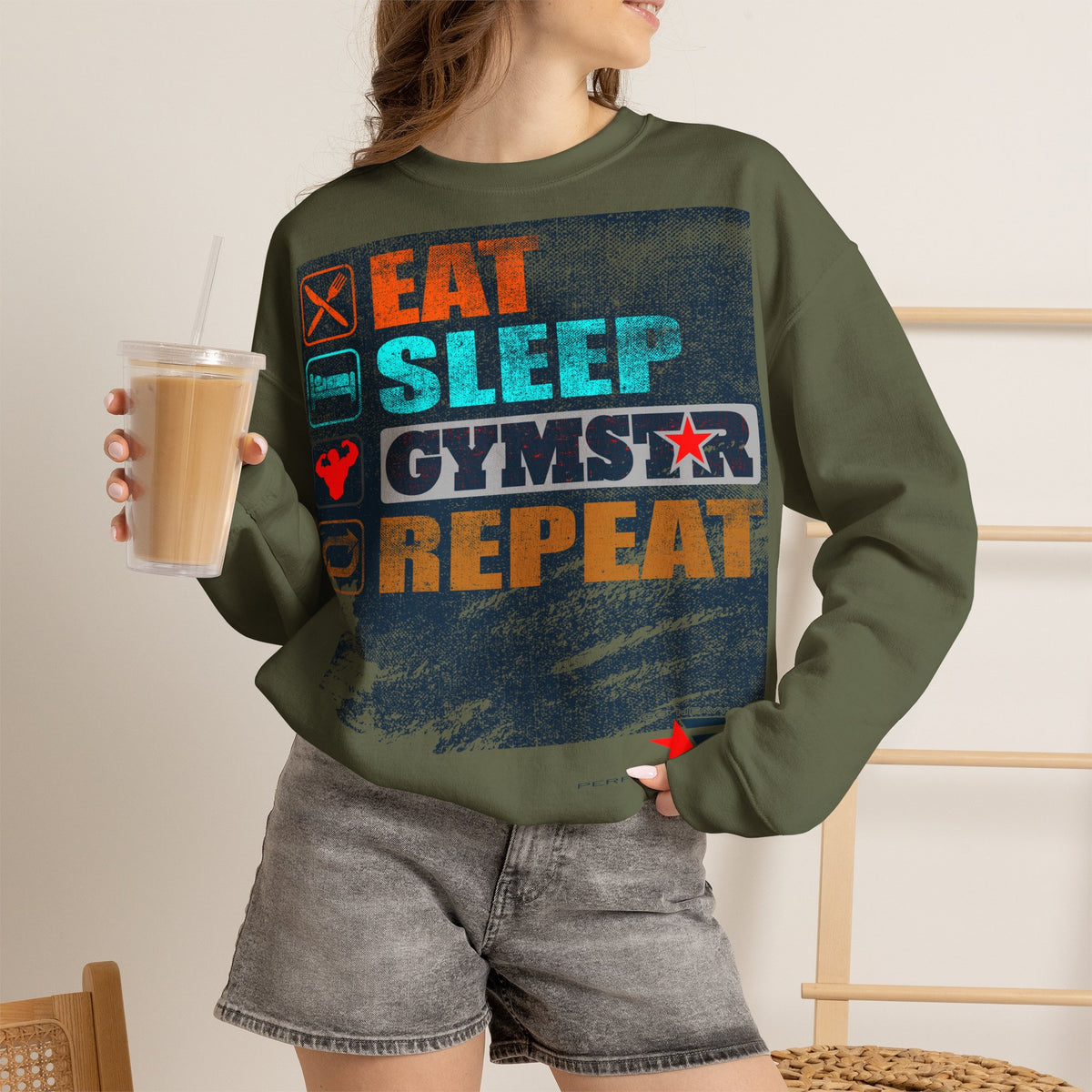 A woman wearing an olive green crewneck sweatshirt with a colorful graphic that reads 'EAT SLEEP GYMSTAR REPEAT' in orange, blue, and white, paired with gray denim shorts and holding a iced coffee.