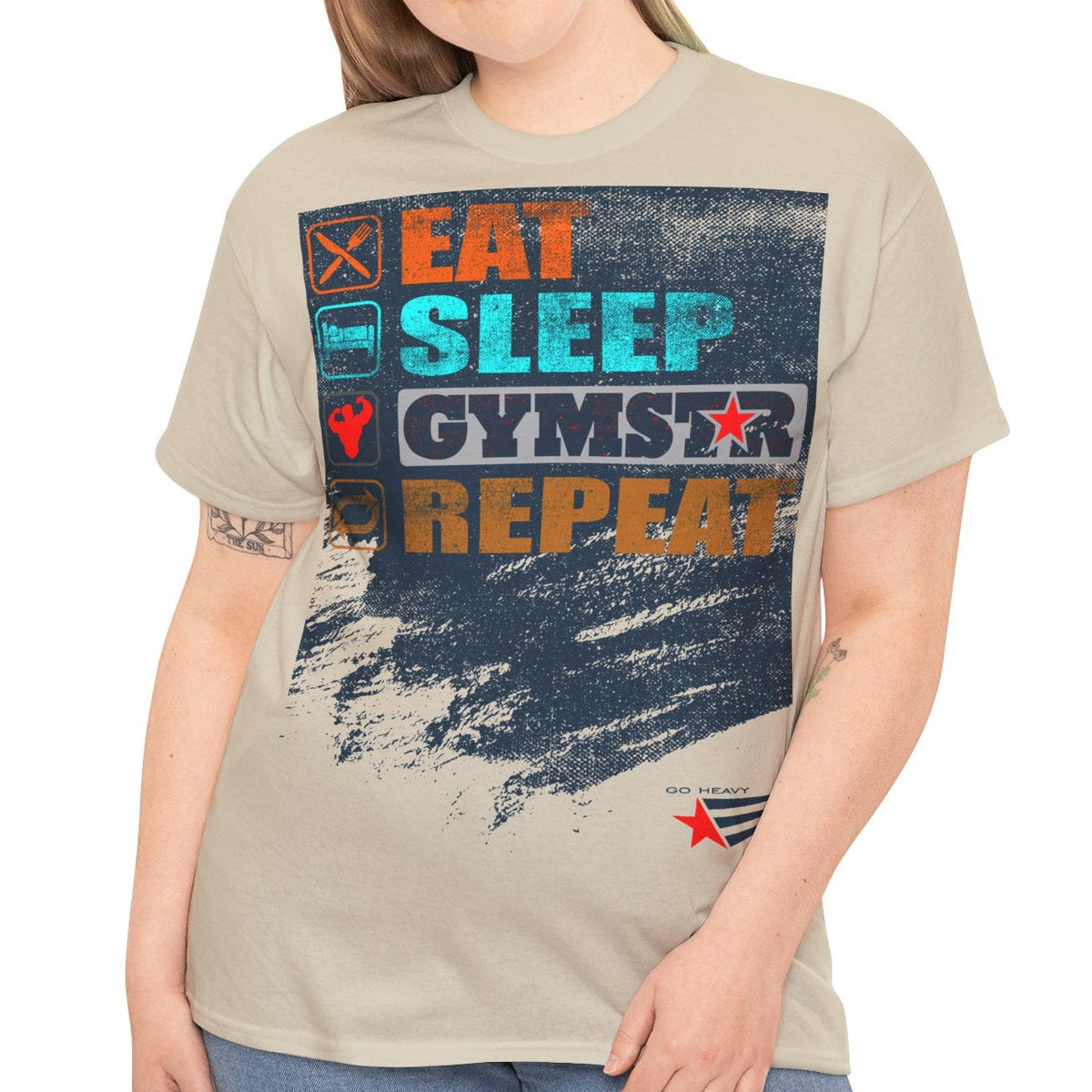A woman modeling a light beige graphic t-shirt with a distressed gym-themed print that reads 'EAT SLEEP GYMSTAR REPEAT' in orange, blue, and white, paired with blue jeans against a plain white background.