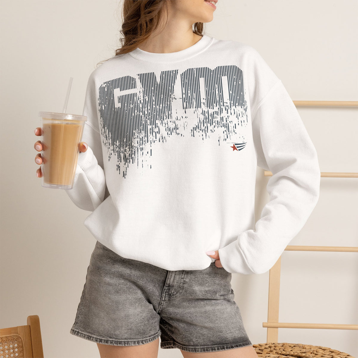 A woman wearing a white crewneck sweatshirt with a large gray pixelated 'GYM' graphic on the front, paired with gray denim shorts and holding an iced coffee.