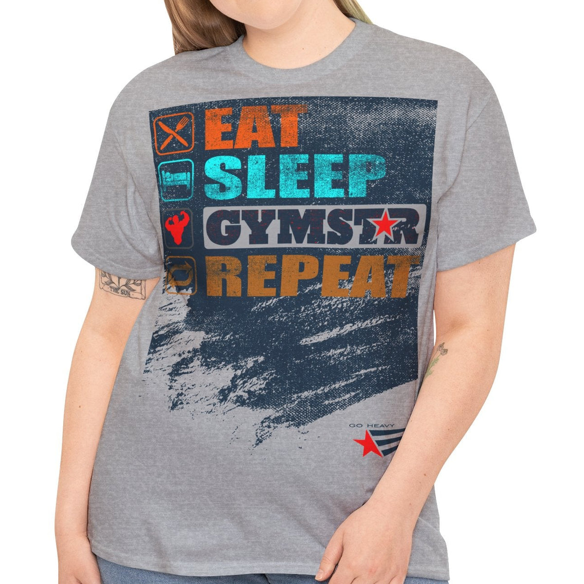 A woman modeling a heather gray graphic t-shirt with a distressed gym-themed print that reads 'EAT SLEEP GYMSTAR REPEAT' in orange, blue, and white, paired with blue jeans against a plain white background.