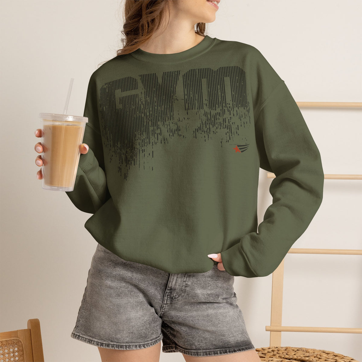 A woman wearing an olive green crewneck sweatshirt with a black distressed 'GYM' graphic on the front, paired with gray denim shorts and holding an iced coffee.