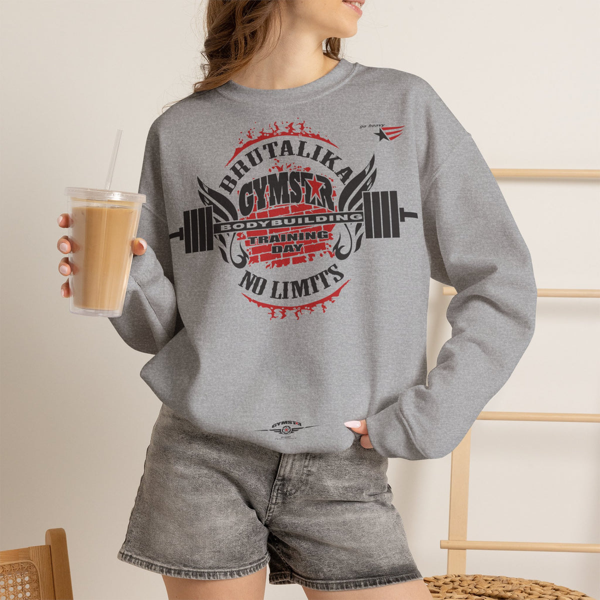 A woman wearing a heather gray crewneck sweatshirt with a red and black 'Brutalika Gym' graphic, paired with gray denim shorts, holding a drink in a warm indoor setting.
