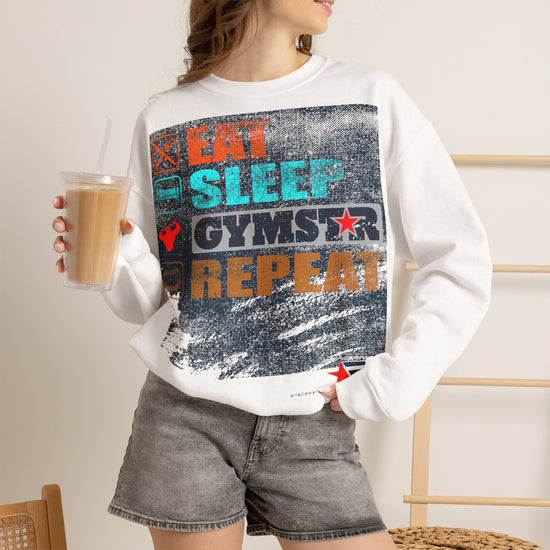 A woman wearing a white crewneck sweatshirt with a colorful distressed graphic that reads 'EAT SLEEP GYMSTAR REPEAT' in orange, teal, and dark blue. She is holding a cup of iced coffee and wearing gray denim shorts.