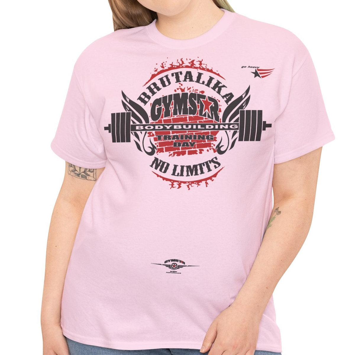 A woman modeling a light pink short-sleeve t-shirt with a large graphic on the front that reads 'BRUTALIKA GYMSTAR NO LIMITS' in red and black, paired with blue jeans, against a plain white background.