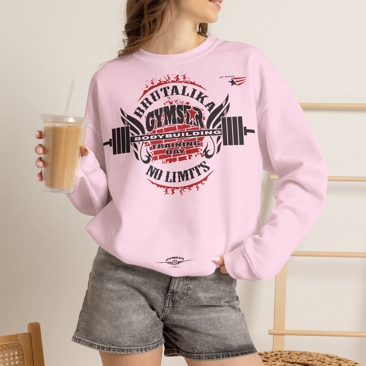 A woman wearing a light pink crewneck sweatshirt with a large red, black, and white graphic on the front that reads 'BRUTALIKA GYMSTAR NO LIMITS' with a barbell design, paired with gray denim shorts and holding an iced coffee.