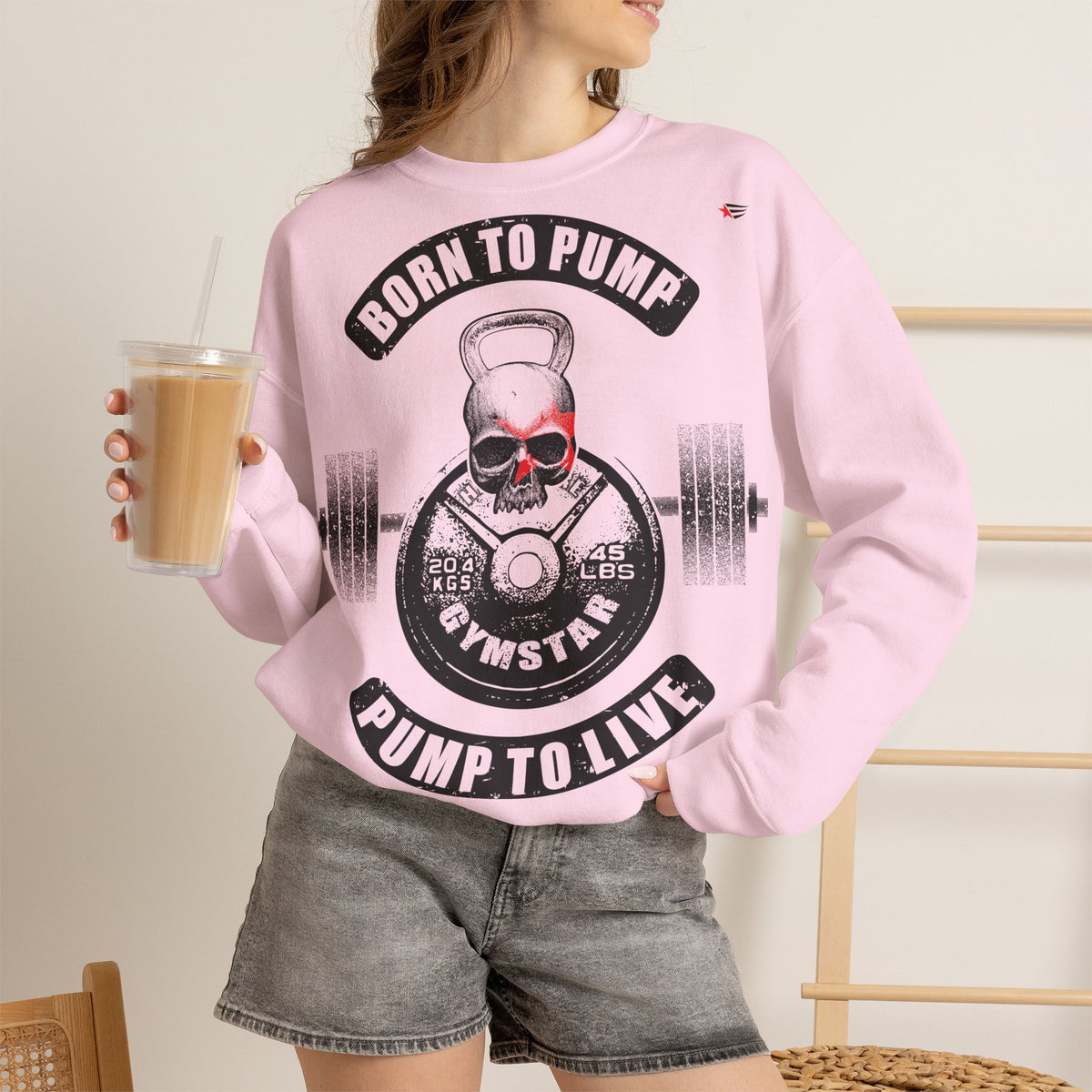 A woman wearing a light pink crewneck sweatshirt with a black and red skull graphic that reads 'BORN TO PUMP PUMP TO LIVE' and 'GYMSTAR'. She is holding an iced coffee and wearing gray denim shorts in a brightly lit indoor setting.