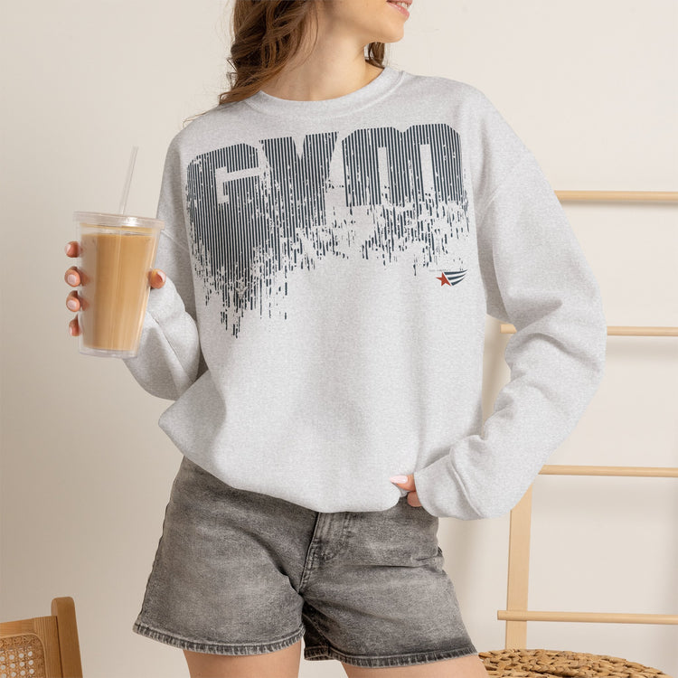 A woman wearing a light gray crewneck sweatshirt with a distressed black 'GYM' graphic and a small red star logo, paired with gray denim shorts and holding an iced coffee.