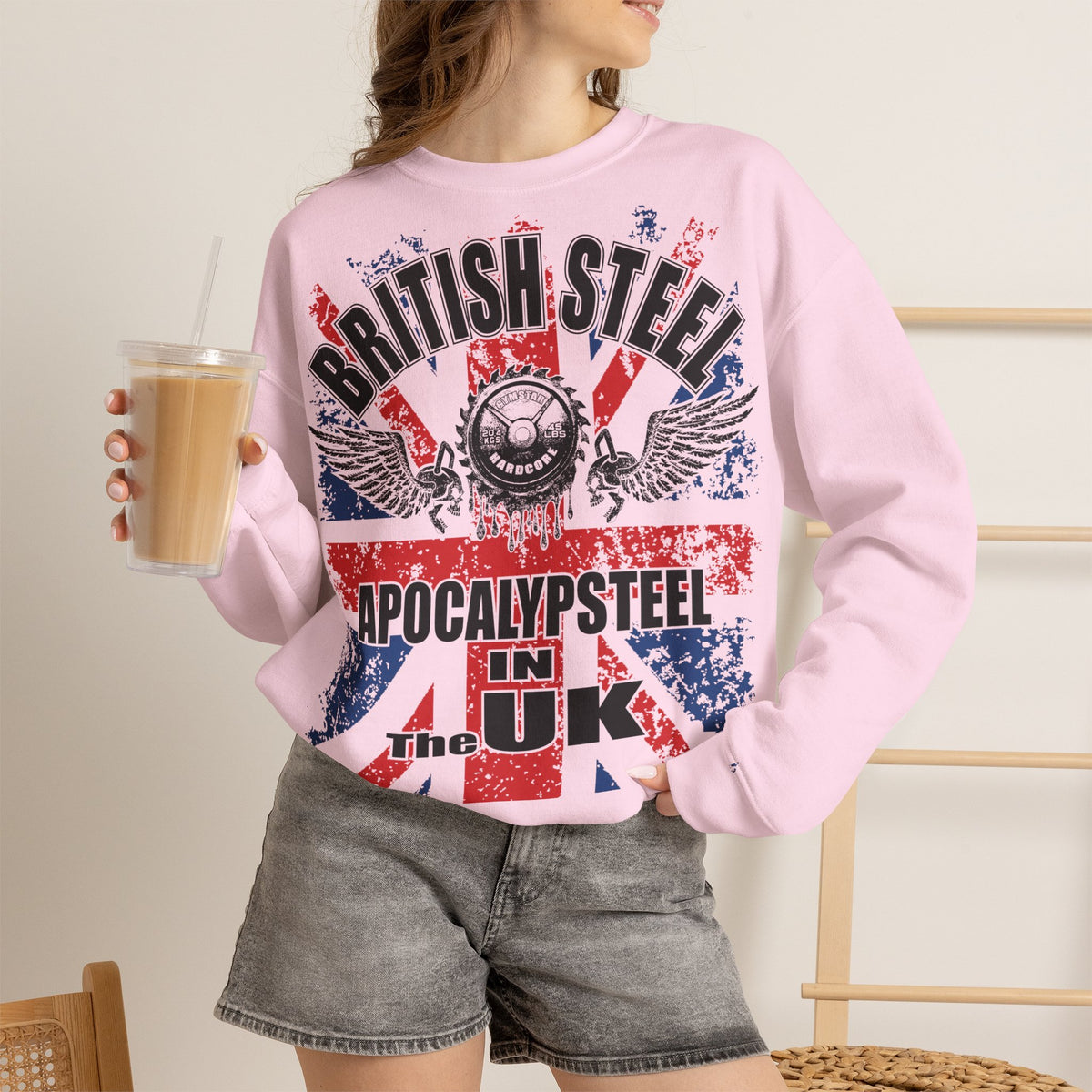 A woman wearing a light pink crewneck sweatshirt with a distressed red, white, and blue graphic that reads 'BRITISH STEEL APOCALYPSTEEL IN UK' and 'The Great Steel of the Gods'. She is holding a cup of iced coffee and wearing gray denim shorts.
