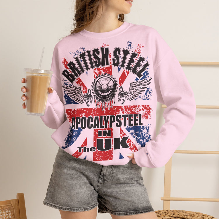 A woman wearing a light pink crewneck sweatshirt with a distressed red, white, and blue graphic that reads 'BRITISH STEEL APOCALYPSTEEL IN UK' and 'The Great Steel of the Gods'. She is holding a cup of iced coffee and wearing gray denim shorts.