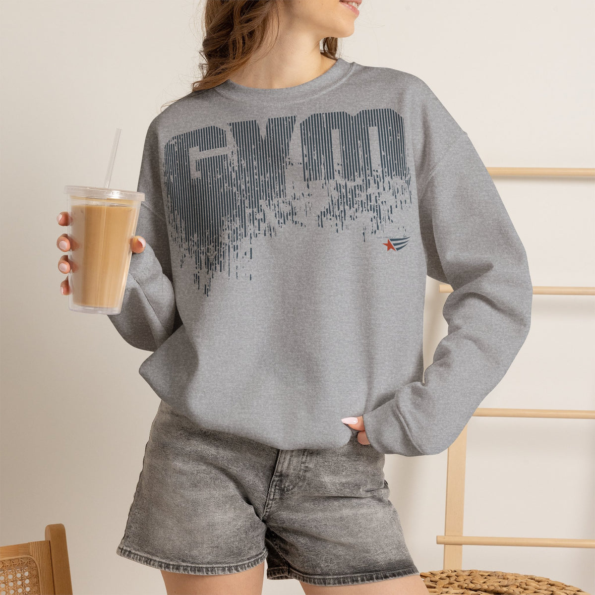 A woman wearing a heather gray crewneck sweatshirt with a dark gray 'GYM' graphic and a small red star logo on the chest, paired with gray denim shorts and holding an iced coffee.