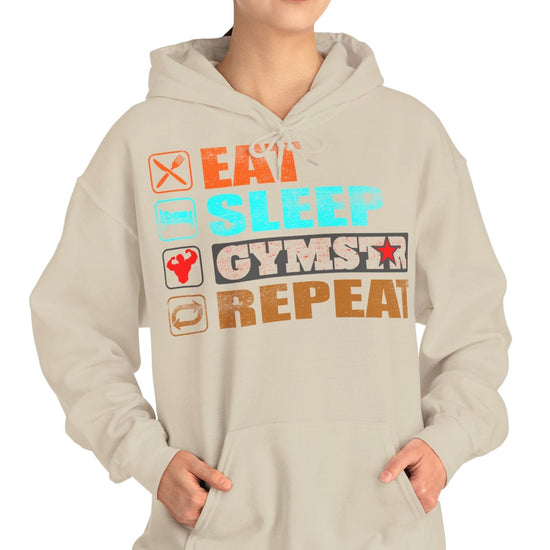 A woman modeling a light beige hoodie with a colorful graphic print that reads 'EAT SLEEP GYMSTAR REPEAT' with small icons, set against a plain white background.