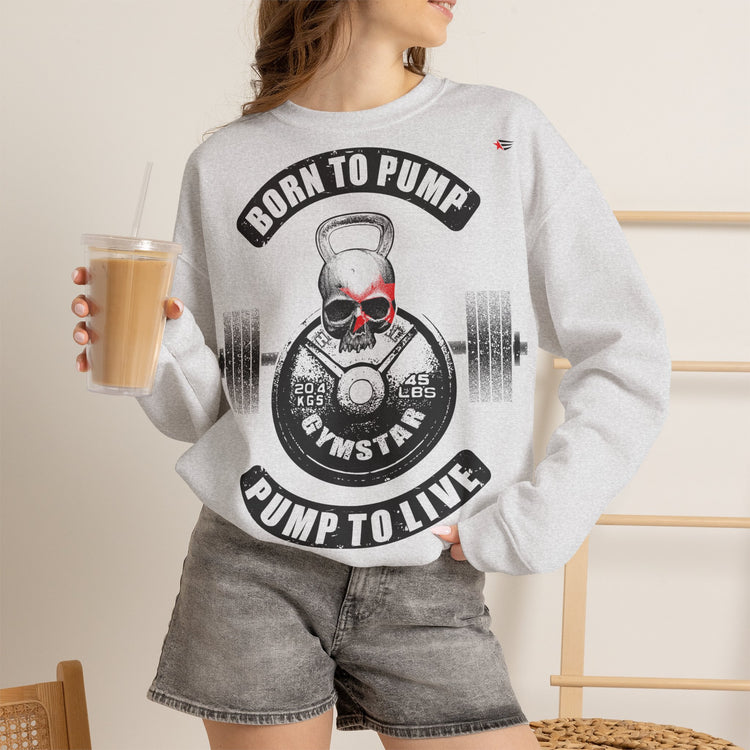 A woman wearing a light gray crewneck sweatshirt with a black and red graphic that reads 'BORN TO PUMP' and 'PUMP TO LIVE' around a skull and barbell design. She is also wearing gray denim shorts and holding an iced coffee in a cafe setting.