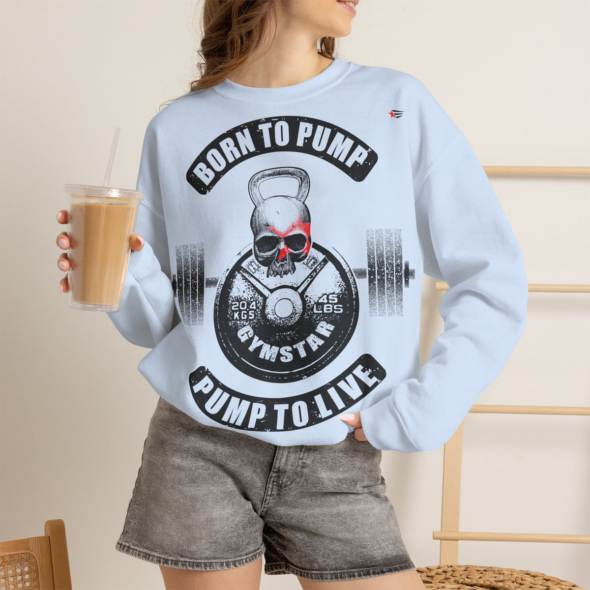 A woman wearing a light blue crewneck sweatshirt with a black and white skull graphic and the text 'BORN TO PUMP PUMP TO LIVE' and 'GYMSTAR'. She is holding an iced coffee in a cafe setting.