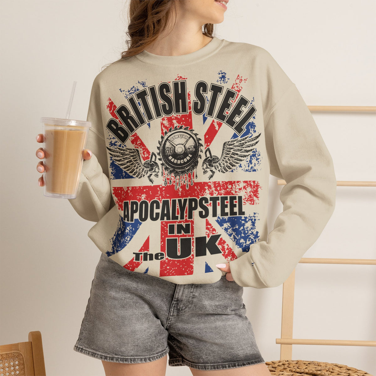 A woman wearing a beige crewneck sweatshirt with a distressed red, white, and blue graphic that reads 'BRITISH STEEL APOCALYPSTEEL IN THE UK', paired with gray denim shorts and holding an iced coffee.
