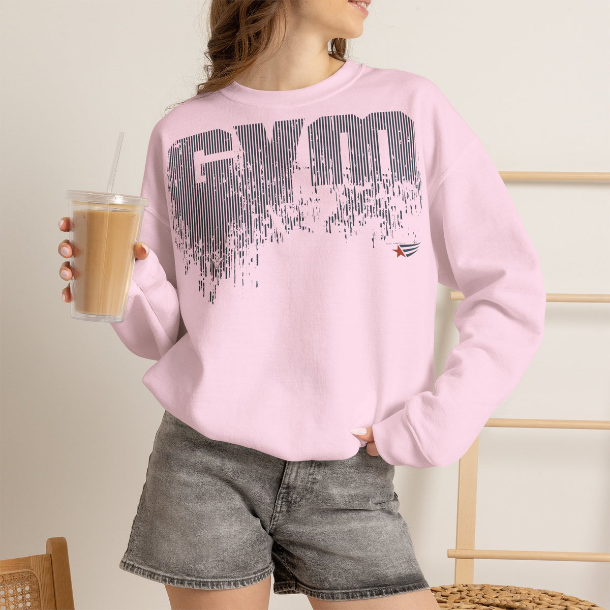 A woman wearing a light pink crewneck sweatshirt with a distressed black 'GYM' graphic on the front, paired with gray denim shorts and holding an iced coffee.