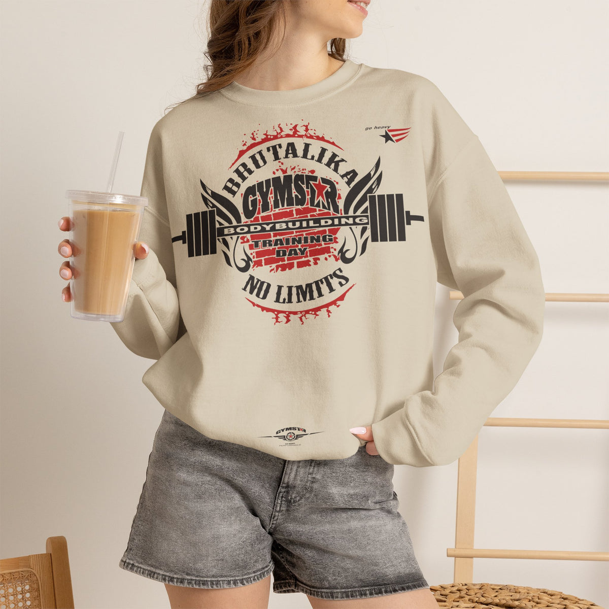 A woman wearing a beige crewneck sweatshirt with a bold red and black gym graphic on the front, paired with gray denim shorts and holding an iced coffee.