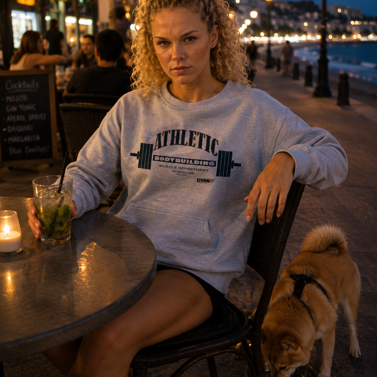Woman wearing grey Gymstar sweatshirt sitting at a table with a dog by her side, both on a waterfront promenade at dusk.