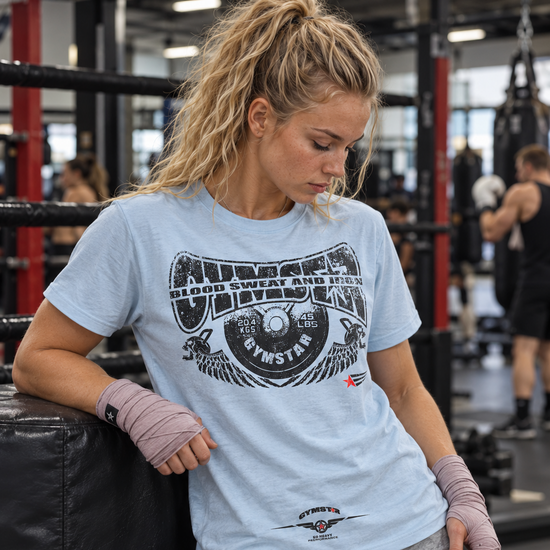 Woman beside a boxing ring wearing a light blue GYMSTAR Blood, Sweat and Iron T-shirt and boxing hand wraps.