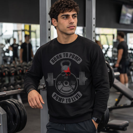 A man in a gym wearing a black long-sleeve graphic sweatshirt with a skull and kettlebell design and the text 'BORN TO PUMP PUMP TO LIVE'.