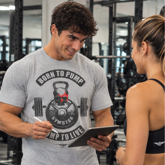 Luca Moretti wearing a sport grey GYMSTAR Born to Pump graphic T-shirt with a kettlebell skull design, smiling while holding a tablet and stylus and talking to a woman in a gym.