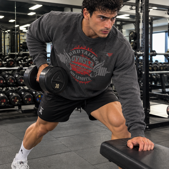 Luca Moretti wearing a dark grey GYMSTAR Brutalika graphic sweatshirt, seated on a gym bench row a dumbbell.