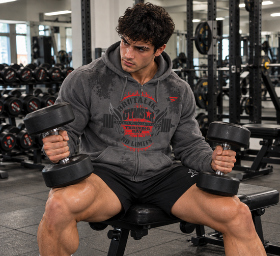 Luca Moretti wearing a grey GYMSTAR Brutalika full-zip hoodie with red and black graphic print, seated on a gym bench holding two dumbbells after an intense workout.