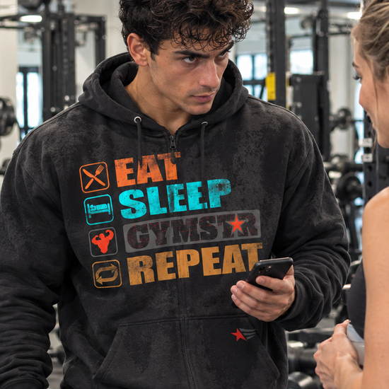 Luca Moretti wearing a black GYMSTAR Eat Sleep Repeat full-zip hoodie, holding a smartphone and workout towel while talking to a woman in a gym.